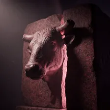Cabeça de touro esculpida em relevo num bloco de granito vermelho escuro, com um dos chifres quebrado, iluminada por uma luz rosa suave que vaza de uma fenda na pedra ao lado