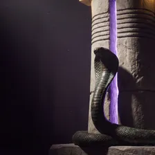 Naja de bronze oxidado erguida junto a um pilar egípcio de pedra com anéis esculpidos, e uma fenda de luz violeta subindo pelo pilar até um toque de luz dourada no alto