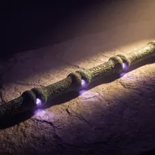Cajado de bronze antigo sobre laje de pedra, com os nós esculpidos ao longo da haste acesos por luz violeta
